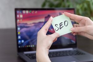 Hands holding SEO note in front of a laptop with a vibrant screen background.